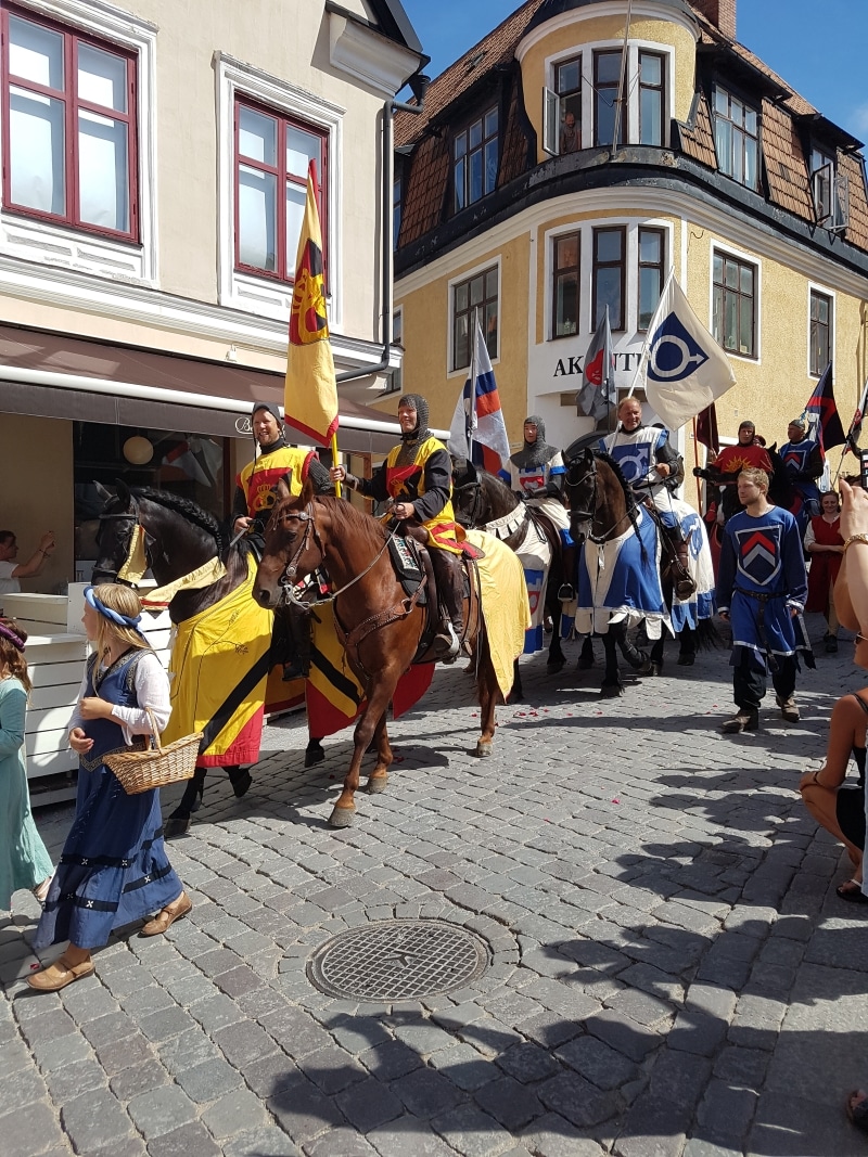 Flera hästar samt människor i medeltidsutstyrsel tågar på kullerstensgatan