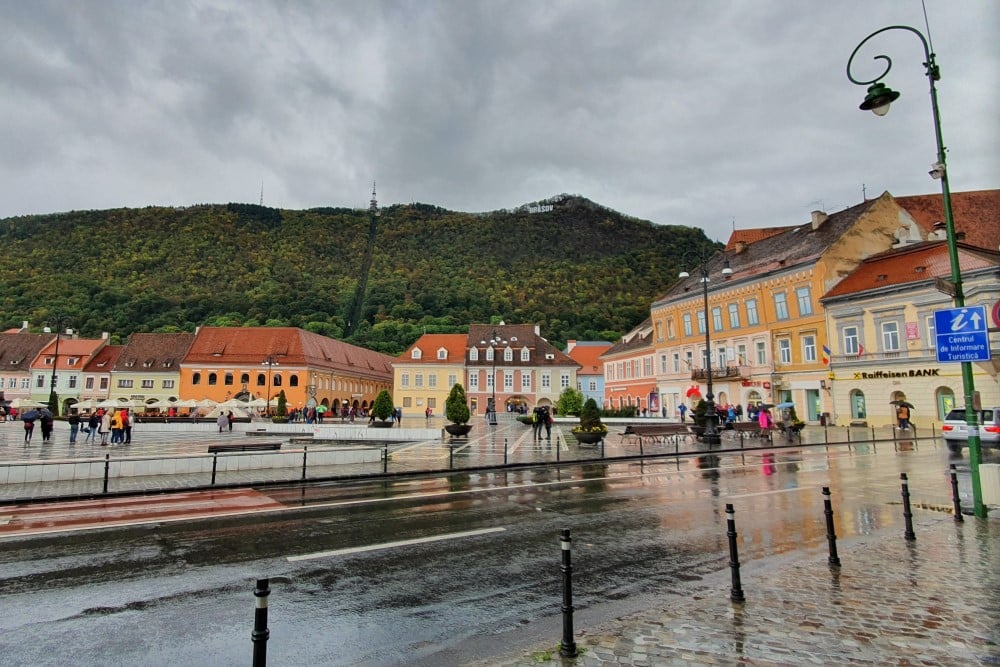 Torg i regnig stad med färgglada byggnader och på håll en kulle med bokstäver som bildar ordet BRASOV