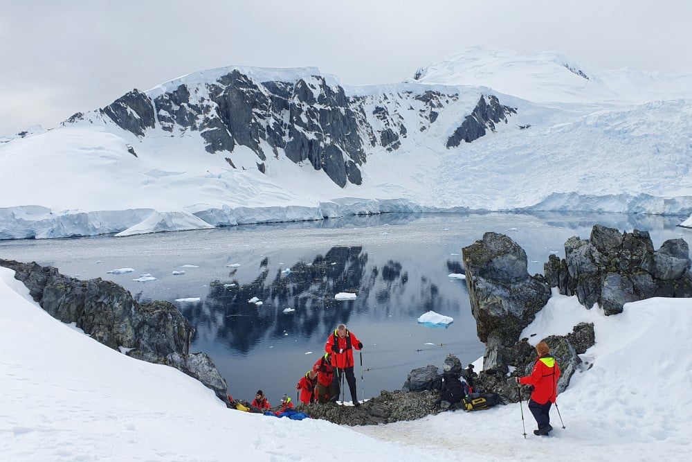 Människor tar sig upp för kulle med utsikt över antarktiska oceanen och snötäckta berg