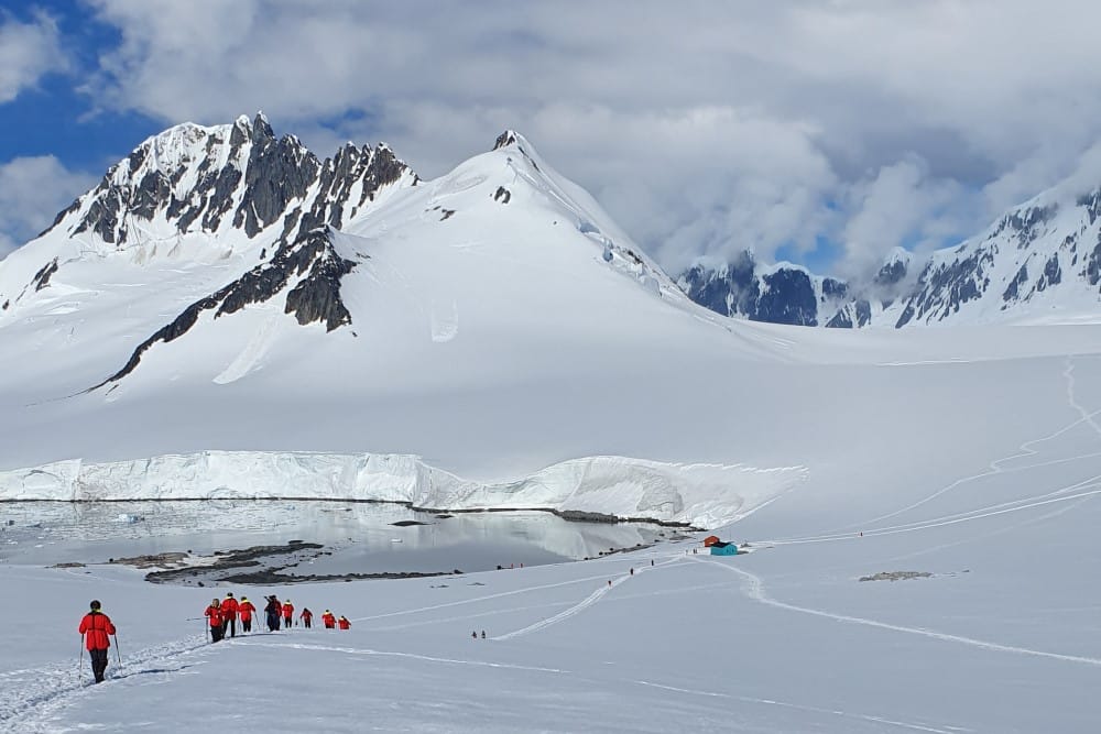 Människor med stavar på promenad i snötäckt mark med berg och delar av antarktiska oceanen