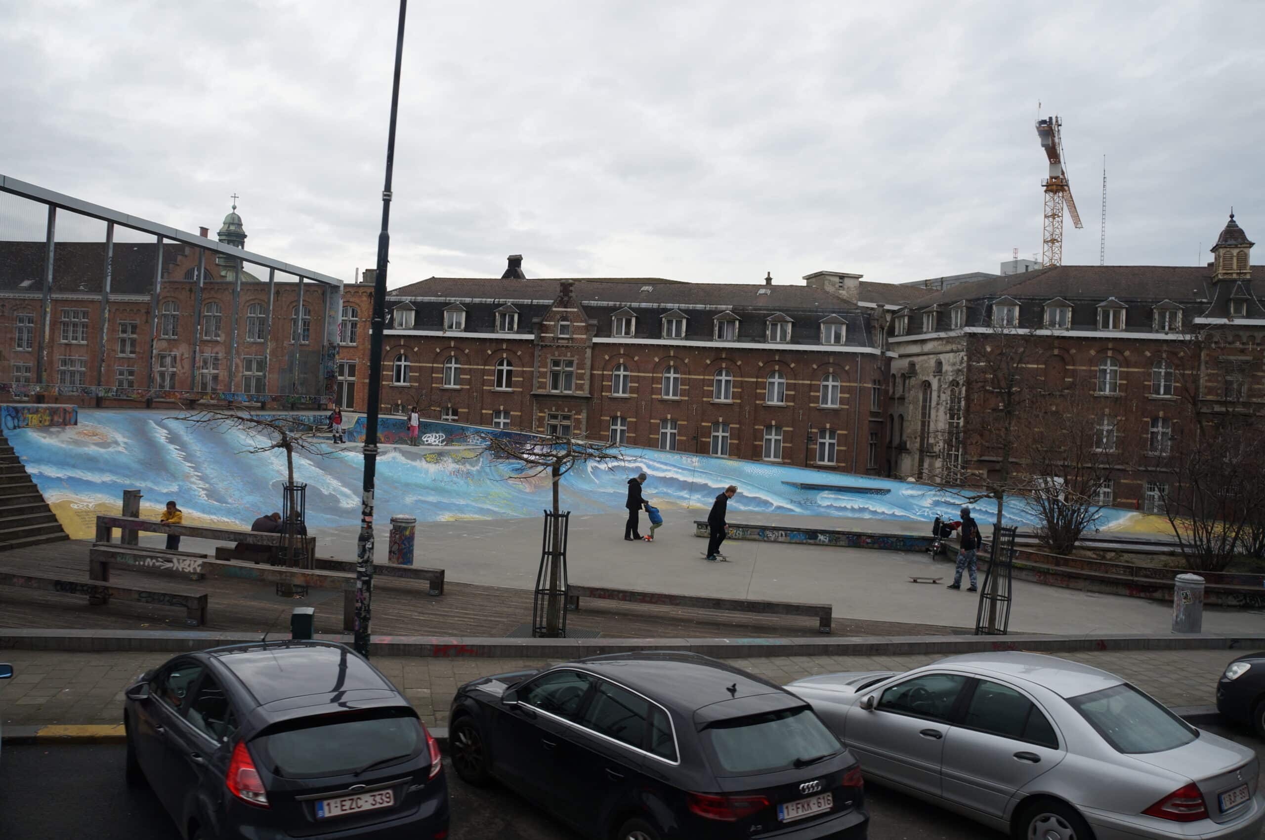 Skatepark målad i blått och gult. Bilar parkerade framför samt bruna byggnader bakom.