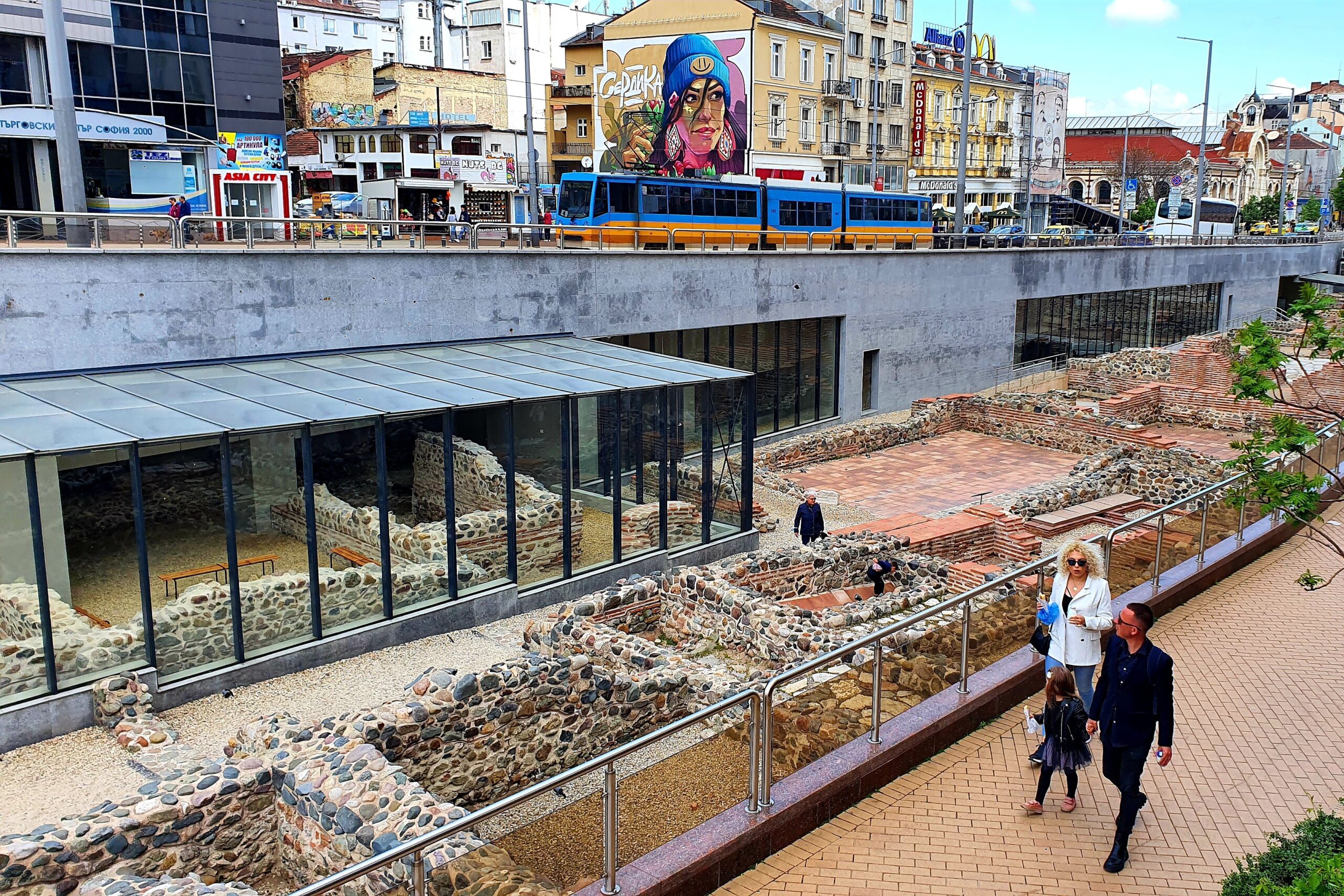 Antika ruiner mitt i stad med byggnader, spårvagnar och människor i rörelse